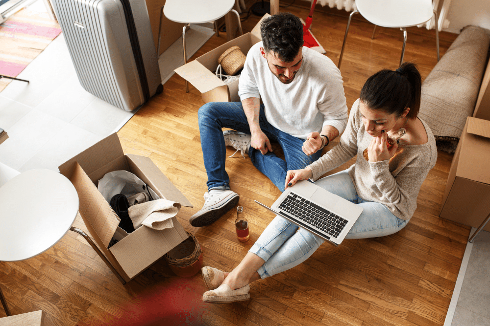 couple sat on the floor surrounded by cardboard boxes on moving day